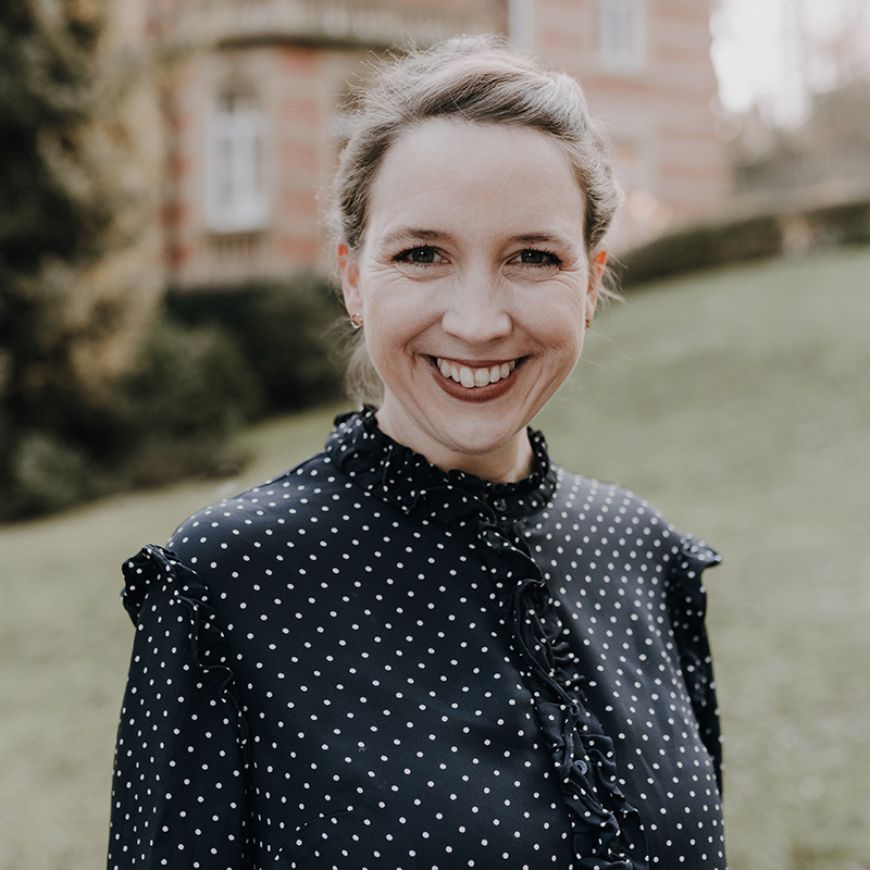 Portrait Eine lächelnde Frau mit zurückgekämmtem hellem Haar trägt eine schwarze Bluse mit weißen Punkten. Sie steht draußen, vielleicht nach einem ihrer Freie-Redner-Workshops auf dem Redner Campus, und im Hintergrund ist ein großes Gebäude mit Bäumen und Sträuchern verschwommen.