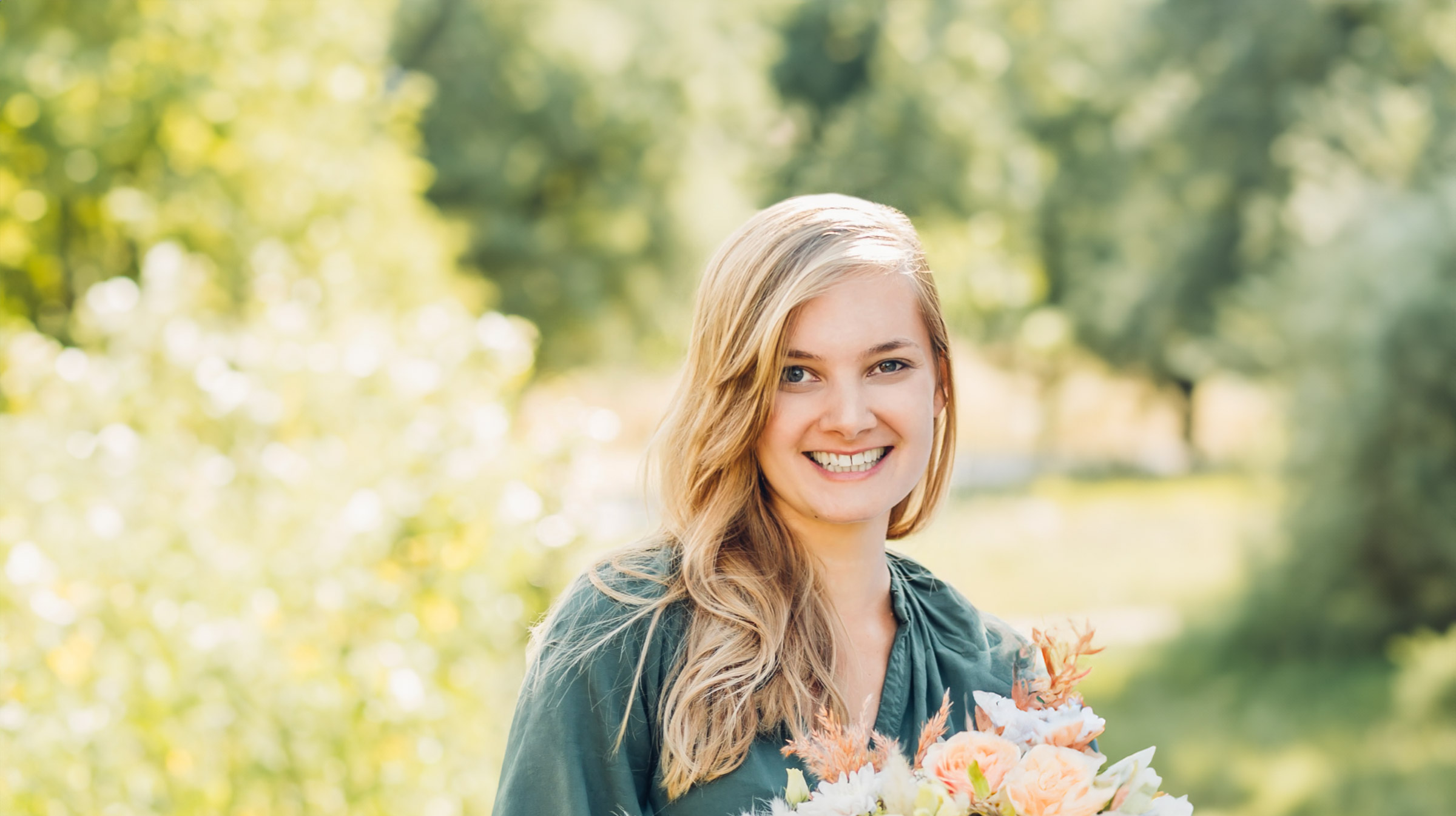 Eine lächelnde Frau mit langen blonden Haaren steht im Freien und hält einen Blumenstrauß in der Hand. Sie trägt ein dunkelgrünes Oberteil und der Hintergrund ist voller Grün, was einen sonnigen Tag in einem üppigen Garten oder Park vermuten lässt, wo sie möglicherweise an Workshops zu Freiem Redner von Redner Campus teilnimmt.