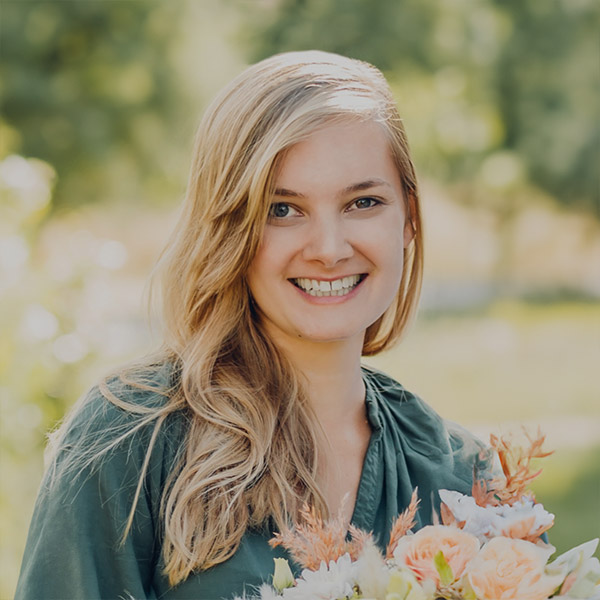 Portrait Eine lächelnde Frau mit langen blonden Haaren steht im Freien und hält einen Blumenstrauß in der Hand. Sie trägt ein dunkelgrünes Oberteil und der Hintergrund ist voller Grün, was einen sonnigen Tag in einem üppigen Garten oder Park vermuten lässt, wo sie möglicherweise an Workshops zu Freiem Redner von Redner Campus teilnimmt.