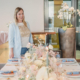 Eine Frau mit blonden Haaren, einem hellblauen Blazer und einem bedruckten T-Shirt, steht neben einem schön gedeckten Esstisch, der mit Blumen und Kerzen dekoriert ist. Im Hintergrund steht eine große Vase mit weißen Orchideen. Der Freie Redner vom Redner Campus lächelt herzlich.