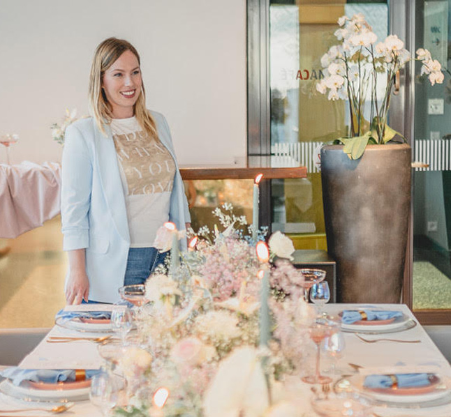 Eine Frau mit blonden Haaren, einem hellblauen Blazer und einem bedruckten T-Shirt, steht neben einem schön gedeckten Esstisch, der mit Blumen und Kerzen dekoriert ist. Im Hintergrund steht eine große Vase mit weißen Orchideen. Der Freie Redner vom Redner Campus lächelt herzlich.