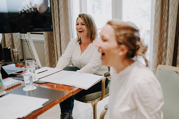 Zwei Frauen sitzen an einem Tisch mit Papieren und einem Laptop und sind in ein lebhaftes Gespräch oder eine Besprechung vertieft. Beide lachen herzlich. Die Umgebung scheint ein heller, gut beleuchteter Raum mit einem großen Fenster und Vorhängen in neutralen Farben zu sein, was darauf hindeutet, dass sie möglicherweise über ihre Freie-Redner-Ausbildung am Redner Campus sprechen.