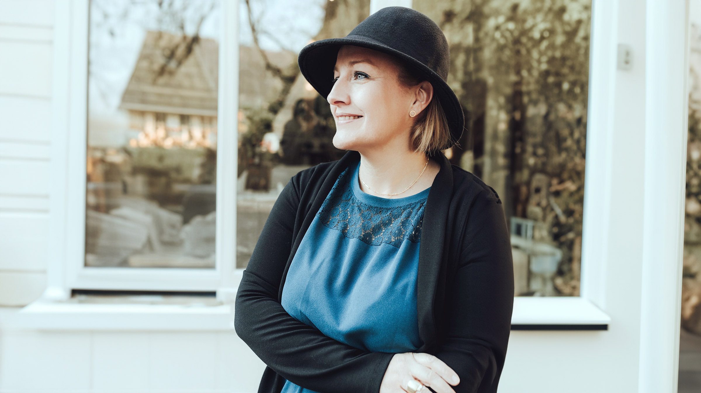 Eine Frau mit dunklem Hut und schwarzer Strickjacke über einem blauen Kleid steht mit verschränkten Armen da, lächelt und blickt nach links. Sie steht vor einem großen Fenster, in dem sich eine Szene im Freien spiegelt, und denkt vielleicht über den bevorstehenden Freie-Redner-Workshop von Redner Campus nach.