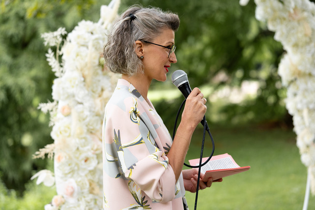 Eine Frau mit grauem Haar und Brille spricht in ein Mikrofon und hält dabei ein rosa Buch mit Notizen in der Hand. Sie steht draußen vor einem mit weißen Blumen und Grünpflanzen geschmückten Bogen. Als Freie Rednerin vom Redner Campus trägt sie eine helle Bluse mit Blumenmuster, die den Menschen um sie herum Trost spendet.