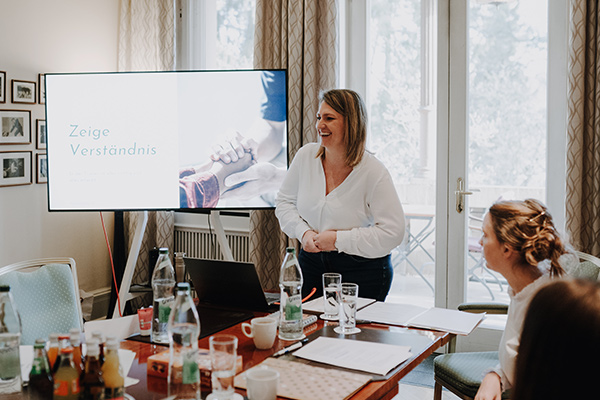 Eine Frau steht und lächelt, während sie vor einer kleinen Gruppe in einem Raum eine Präsentation über das Freie Redner-Training hält. Auf einem Bildschirm erscheint der Text „Zeige Verständnis“. Zwei Personen sitzen an einem Tisch und schauen der Vortragenden zu. Vor ihnen stehen verschiedene Getränke und Unterlagen. Das Training wird von Redner Campus durchgeführt.