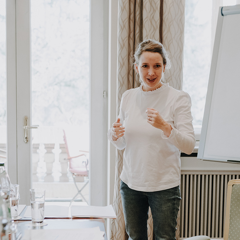 Eine Frau in weißer Bluse steht vor einem Flipchart, spricht und gestikuliert mit ihren Händen. Sie leitet eine Redner Campus-Sitzung für angehende Freie Redner in einem gut beleuchteten Raum mit einem Fenster und einem Balkon im Hintergrund. Wasserflaschen und Papiere sind ordentlich auf dem Tisch neben ihr angeordnet.