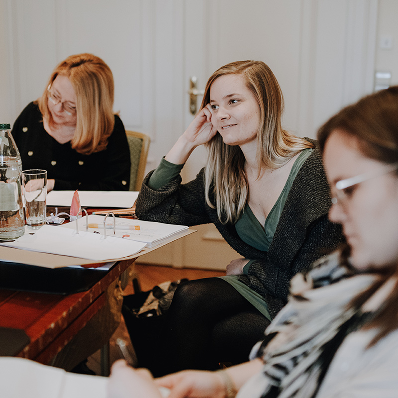 Auschnitt: Drei Frauen sitzen an einem Holztisch und sind offenbar in eine Besprechung oder Diskussion über ihren Freien Redner vertieft. Auf dem Tisch liegen Papiere, Ordner und Getränke. Eine Frau sieht aufmerksam aus, während eine andere wie eine erfahrene Traurednerin vom Redner Campus schreibt und eine dritte Dokumente durchliest.