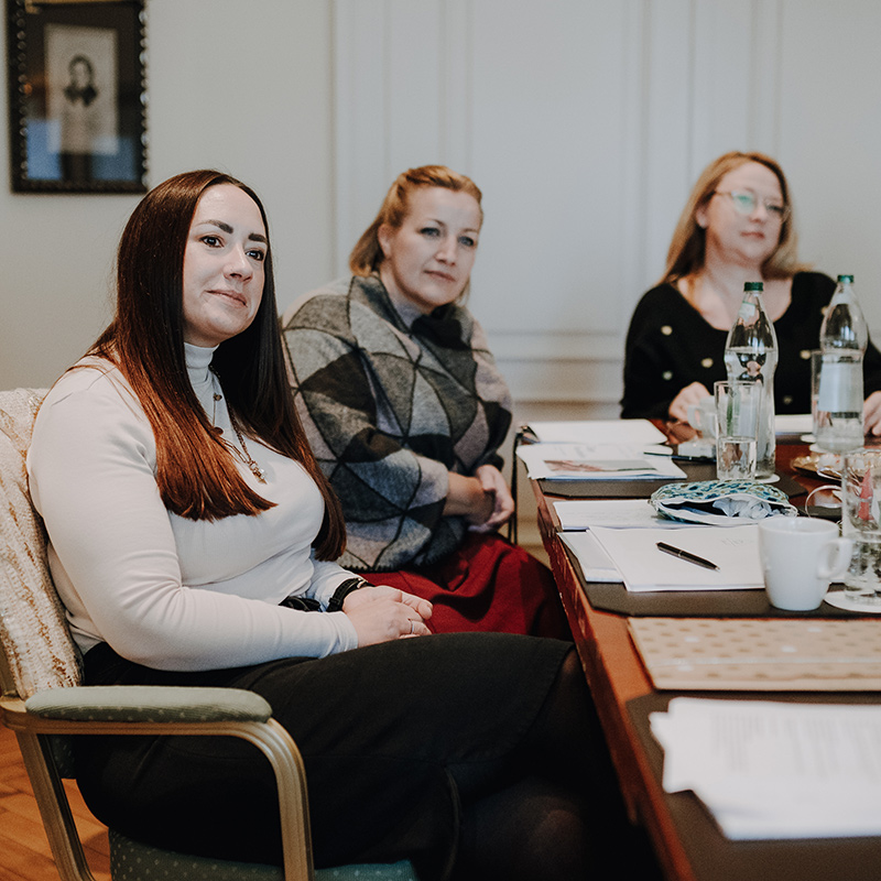 Ausschnit: Drei Frauen sitzen an einem Tisch in einem Besprechungsraum und diskutieren. Der Tisch ist mit Papieren, Wasserflaschen und verschiedenen Getränken aus der Linie „Freie Redner“ des Redner Campus bedeckt. Im Hintergrund hängen gerahmte Porträts an der Wand. Die Frauen wirken aufmerksam und scheinen jemandem aus dem Off zuzuhören, wahrscheinlich während eines ihrer Workshops.