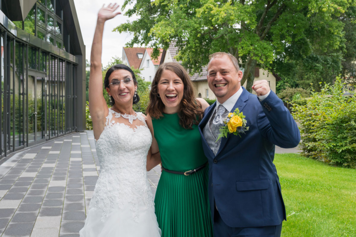 Eine glückliche Braut in einem weißen Kleid und ein Bräutigam in einem blauen Anzug mit einem gelben Blumenboutonniere stehen nach einer Zeremonie im Freien. Die Braut hebt einen Arm und sie lächeln einer Frau in einem grünen Kleid zu, die ihren Arm um sie gelegt hat; es sieht so aus, als wären sie bereit für ihr nächstes großes Abenteuer, vielleicht sogar für gemeinsame Workshops von Frei Redner by Redner Campus.