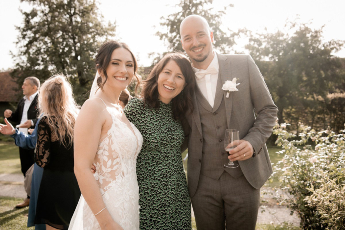 Eine lächelnde Braut in einem weißen Spitzenhochzeitskleid, eine Frau in einem grün gemusterten Kleid und ein Bräutigam in einem grauen Anzug mit weißer Fliege stehen zusammen im Freien. Der Bräutigam hält ein Champagnerglas. Im Hintergrund sind weitere Personen und das Freie Redner des Redner Campus sowie Bäume und Grünflächen zu sehen.