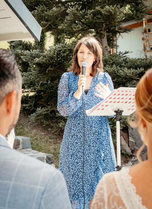 Eine Frau in einem blau gemusterten Kleid spricht bei einer Veranstaltung im Freien, möglicherweise einer Hochzeit, in ein Mikrofon, wie das Paar andeutet, wobei die Frau in einem weißen Kleid von hinten zu sehen ist. Sie steht neben einem Notenständer mit einem Waldstück im Hintergrund, was eine Atmosphäre erzeugt, die an Sitzungen von Frei Redner oder Redner Campus erinnert.